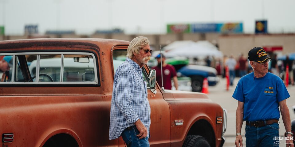 Two Attendees of the 2024 Texas C10 Nationals Stand Outside an Orange Classic Chevrolet Truck