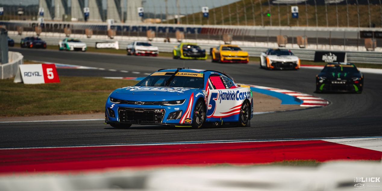 Three Quarters View of a Blue Chevy Racecar Driving Down the Racetrack with Other Racecars Following Suit