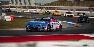 Three Quarters View of a Blue Chevy Racecar Driving Down the Racetrack with Other Racecars Following Suit