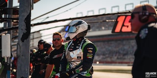 Close View of a Racer wearning a Helmet Sitting Down in Front of the Stadium with People around Him