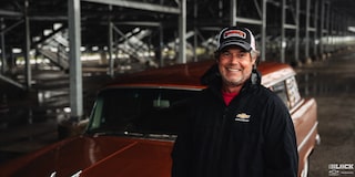 A Man Smiling While Wearing a Cap and a Black Hoodie and Standing in Front of a 1955 Chevy Handyman Wagon