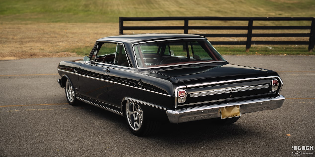 Three Quarters Rear View of a Black 1964 Chevy Nova on an Empty Road in Front of a Grassy Plane